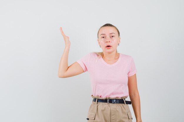 Blond meisje verhogen hand in t-shirt, broek en op zoek in verwarring. vooraanzicht.