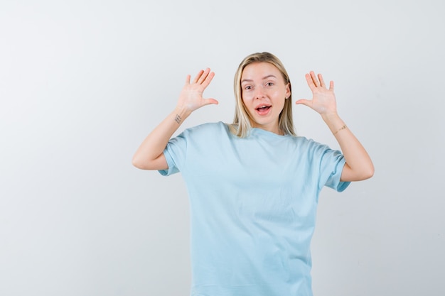 Blond meisje verhogen palmen in overgave gebaar in blauw t-shirt en op zoek verrast, vooraanzicht.