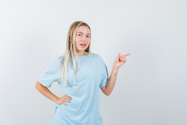 Blond meisje wijst naar rechts met wijsvinger, hand op taille in blauw t-shirt vast te houden en ziet er mooi uit, vooraanzicht.