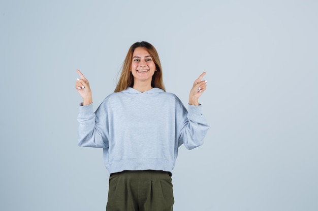 Blond meisje wijst tegenovergestelde richtingen met wijsvinger in olijfgroen blauw sweatshirt en broek en ziet er charmant uit. vooraanzicht.