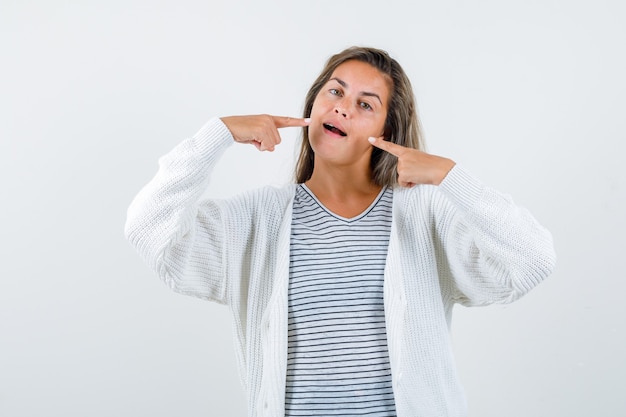 Blond meisje wijzend op tanden in gestreept t-shirt, wit vest en jean broek en ziet er verleidelijk uit. vooraanzicht.