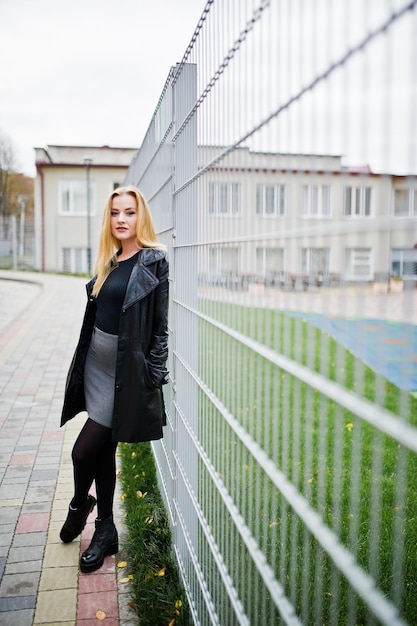 Blond modieus meisje in lange zwarte leren jas tegen ijzeren hek op straat