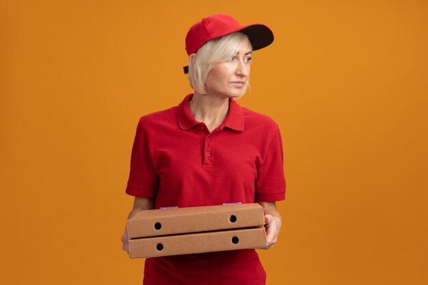 Blonde bezorger van middelbare leeftijd in rood uniform en pet met pizzapakketten kijkend naar kant geïsoleerd op oranje muur met kopieerruimte copy