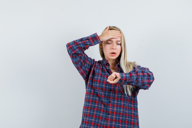 Blonde dame beweert horloge om haar pols in casual overhemd te kijken en vergeetachtig, vooraanzicht te kijken.