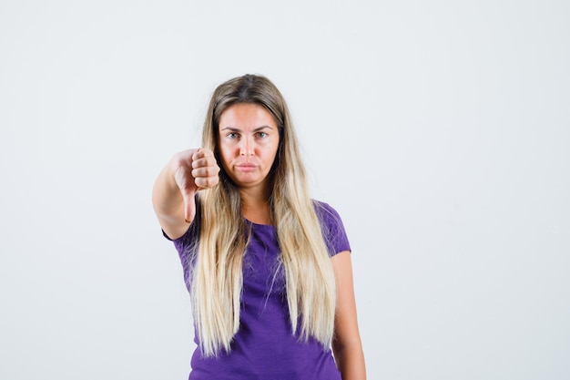 Blonde dame die duim neer in violet t-shirt toont en resoluut, vooraanzicht kijkt.