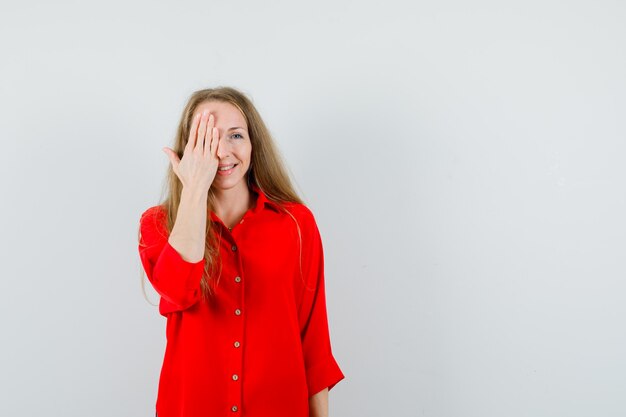 Blonde dame die één oog bedekt met hand in een rood shirt en er opgewonden uitziet,