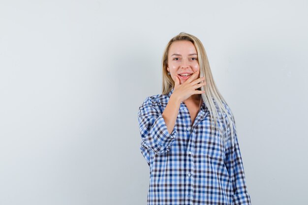 Blonde dame die hand op kin in geruit overhemd houdt en vrolijk kijkt