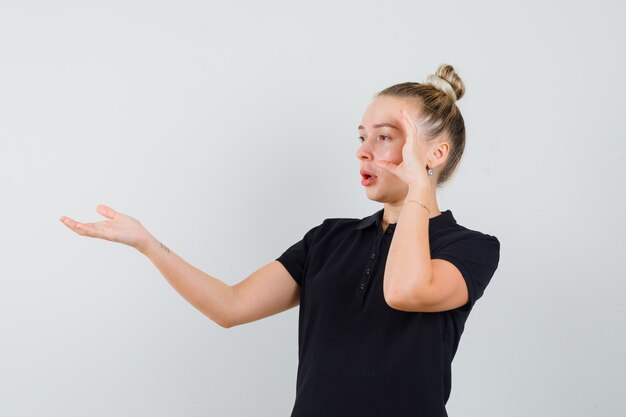 Blonde dame die hand op vragende wijze in zwart t-shirt uitrekt en verbaasd kijkt. vooraanzicht.