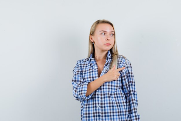 Blonde dame die naar de rechterbovenhoek in een casual overhemd wijst en gefocust kijkt