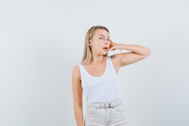 Blonde dame die telefoongebaar in singlet, broek toont en zelfverzekerd, vooraanzicht kijkt.