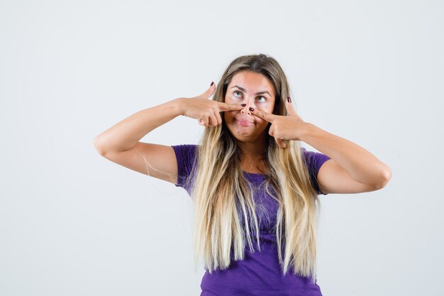 Blonde dame die vingers op neus in violet t-shirt houdt en er grappig uitziet. vooraanzicht.