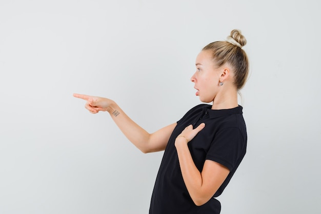 Blonde dame die weg wijst in zwart t-shirt en verbaasd kijkt.