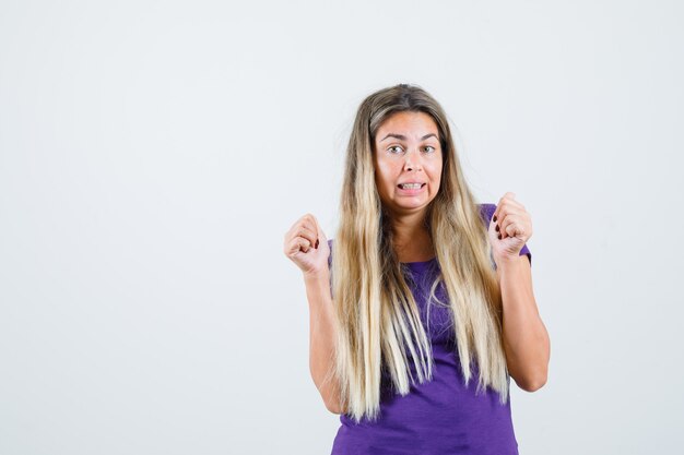 Blonde dame die winnaargebaar in violet t-shirt toont en gelukkig kijkt. vooraanzicht.