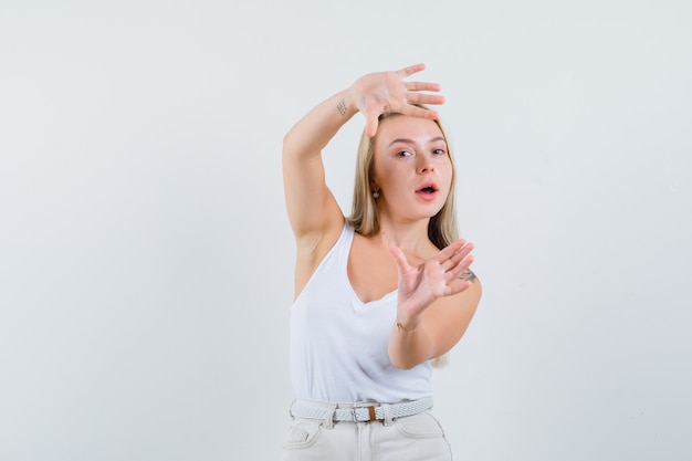 Blonde dame frame gebaar maken in singlet, broek en op zoek naar zelfverzekerd, vooraanzicht.