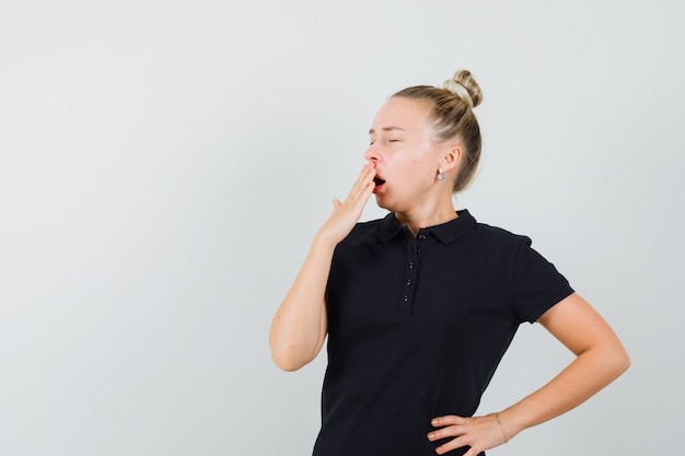 Blonde dame geeuwen in zwart t-shirt en op zoek slaperig, vooraanzicht.