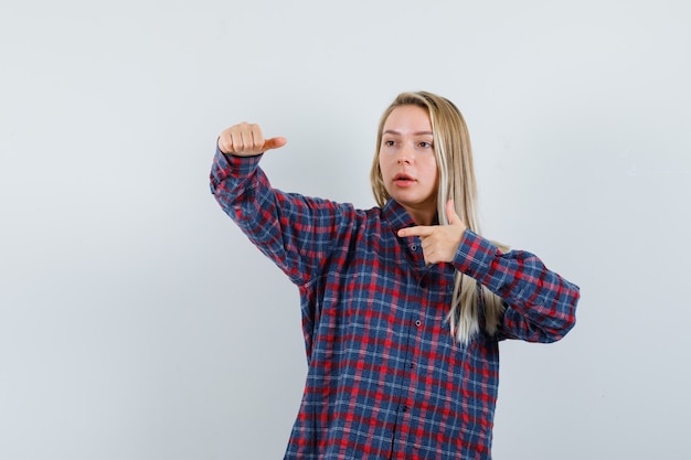 Blonde dame in casual overhemd met duim midden, opzij wijzend en op zoek gericht, vooraanzicht.