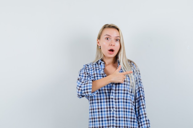 Blonde dame in geruit overhemd wijst naar rechts en kijkt verbaasd