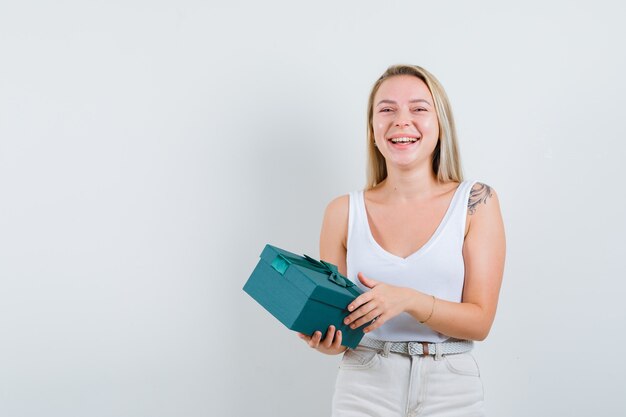 Blonde dame in hemd, broek die huidige doos vasthoudt en er vrolijk uitziet, vooraanzicht.