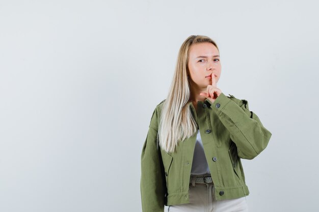 Blonde dame in jasje, broek die stilte gebaar toont en voorzichtig, vooraanzicht kijkt.