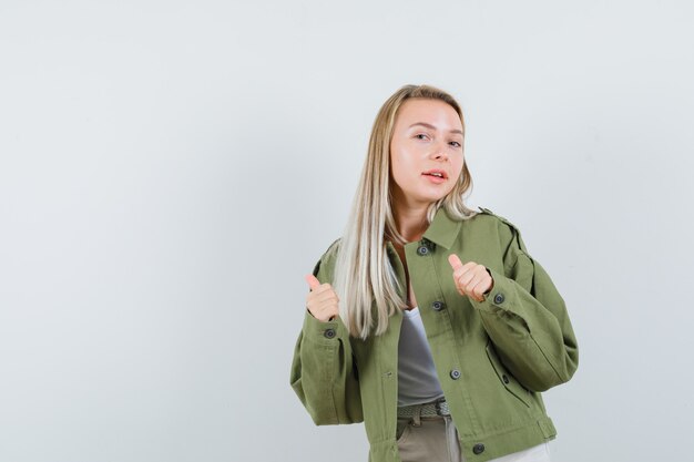 Blonde dame in jasje, broek wijst opzij met duimen omhoog en ziet er zelfverzekerd uit, vooraanzicht.