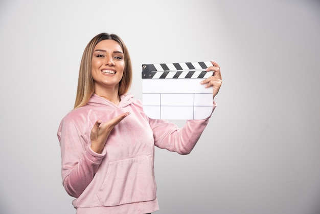 Blonde dame in roze sweatshier met een leeg klepelbord en geeft natuurlijke poses.