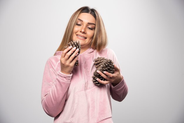 Blonde dame in roze sweatshirt trekt een positief en blij gezicht door eikenhouten kegels vast te houden.