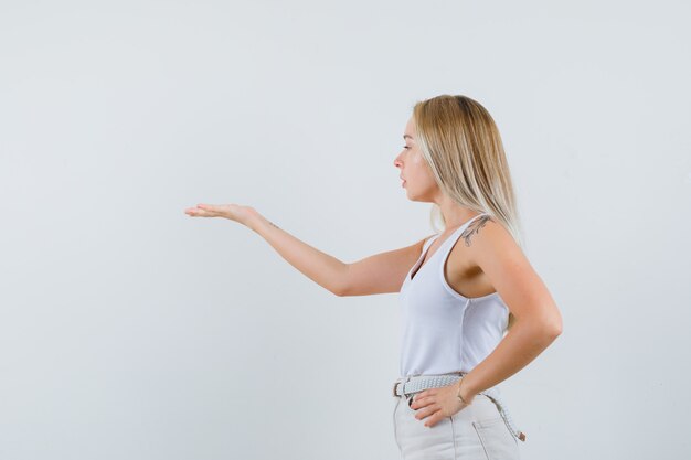 Blonde dame in singlet, broek die hand uitrekt en gericht kijkt.