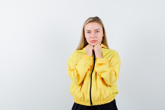 Blonde dame in trainingspak hand in hand onder de kin en op zoek verstandig, vooraanzicht.