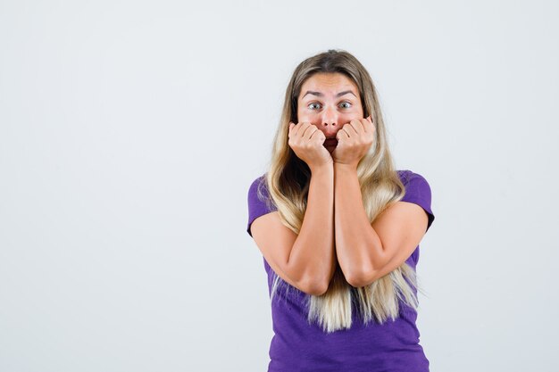 Blonde dame in violet t-shirt vuisten op gezicht te drukken en bang, vooraanzicht op zoek.