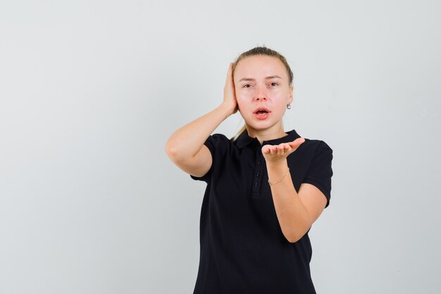 Blonde dame in zwart t-shirt uitrekken hand in verbaasd gebaar, vooraanzicht.