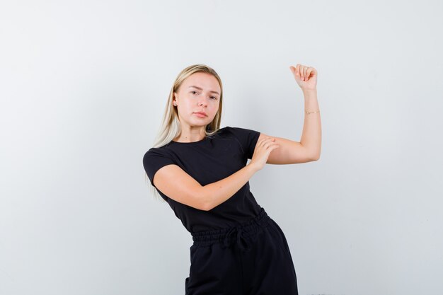 Blonde dame in zwarte jurk met spieren van de arm en op zoek naar zelfverzekerd, vooraanzicht.