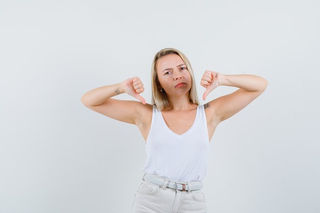 Blonde dame met dubbele duimen naar beneden in hemd, broek en ontevreden, vooraanzicht.