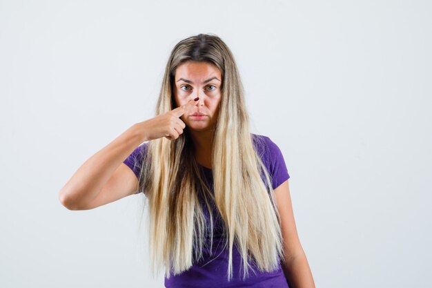 Blonde dame neus met vinger in violet t-shirt, vooraanzicht aan te raken.