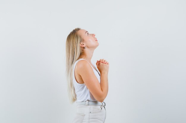 Blonde dame omklemde handen in biddend gebaar in hemd, broek en op zoek hoopvol.