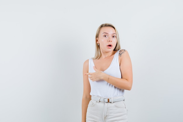 Blonde dame wijst naar de linkerkant in hemd, broek en kijkt verbaasd, vooraanzicht.