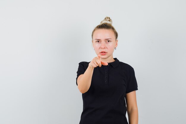 Blonde dame wijst naar voren in zwart t-shirt en kijkt serieus, vooraanzicht.