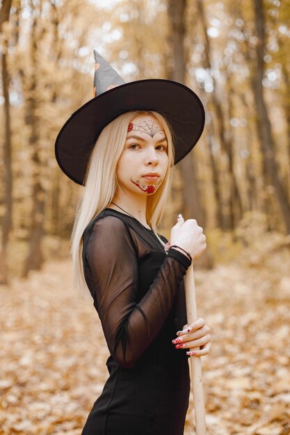 Blonde jonge meisjesheks in bos op Halloween. Meisje met zwarte jurk en kegelhoed. Heks die een bezem vasthoudt.