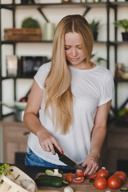 Blonde jonge vrouw die rode tomaten met scherp mes snijdt