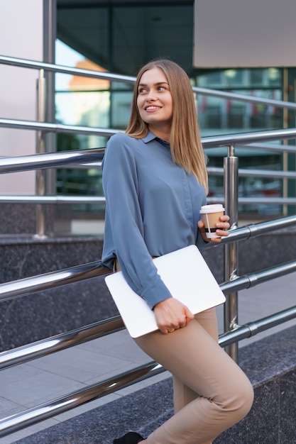 Blonde jonge vrouw lachend portret met laptop en koffie, blauwe zachte shirt dragen over modern gebouw achtergrond