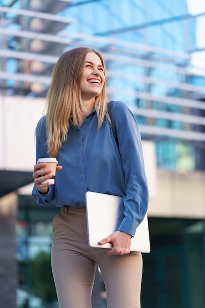 Gratis foto blonde jonge vrouw lachend portret met laptop en koffie, blauwe zachte shirt dragen over modern gebouw