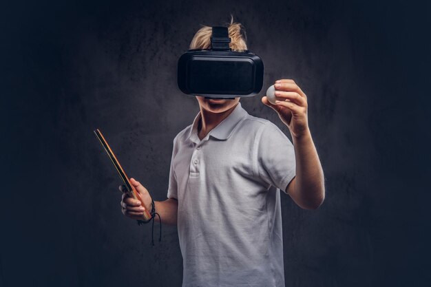 Blonde jongen gekleed in een wit t-shirt die pingpong speelt met een virtual reality-bril. Geïsoleerd op een donkere gestructureerde achtergrond.