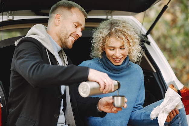 Blonde krullende vrouw en man zitten in een kofferbak in de auto in het herfstbos met een thermokop