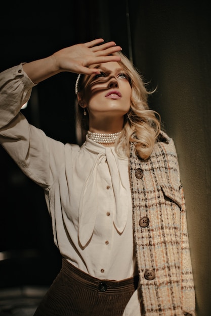 Blonde krullende vrouw in tweed jasje en witte elegante blouse bedekt zon met hand. charmant meisje kijkt op en poseert in een donkere kamer