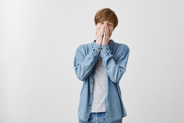 Blonde mannelijke student gekleed in een spijkerbroekoverhemd dat het gezicht bedekt met handen, starend met een verbijsterde blik, moe van stressvol werk, bezorgd over belangrijk examen op de universiteit