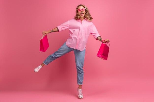 Blonde mooie vrouw in roze shirt en spijkerbroek glimlachend springen op roze achtergrond geïsoleerd met plezier met boodschappentassen