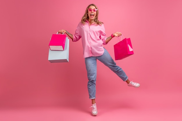 Blonde mooie vrouw in roze shirt en spijkerbroek glimlachend springen op roze geïsoleerd met plezier met boodschappentassen