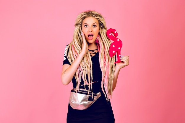 Blonde mooie vrouw met lange blonde dreadlocks met plastic flamingo, stijlvolle sport chique outfit, verrast emoties, pop laatste stijl dragen.