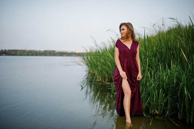 Blonde sensuele vrouw in rode marsala-jurk die in het water van het meer met riet staat