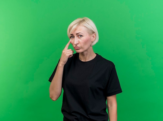 Blonde Slavische vrouw op middelbare leeftijd die voorzijde bekijkt die vinger onder oog zet die op groene muur met exemplaarruimte wordt geïsoleerd