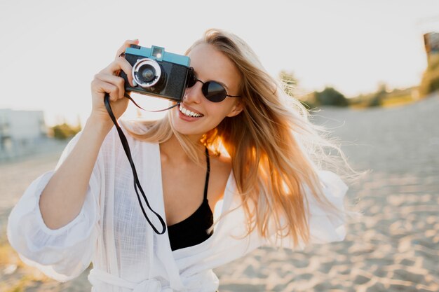 Blonde speelse vrouw die retro camera houdt en pret op warm zonnig strand heeft. Zomervakantie en reizen concept. Natuurlijke schoonheid, vakantie in Azië. Trendy zonnebril, witte outfit.